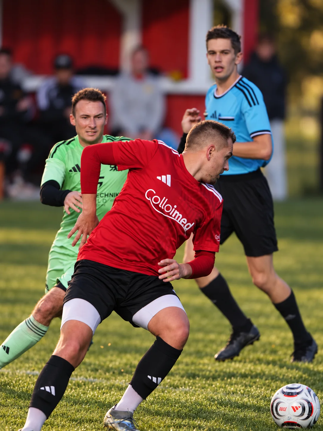 Fußballspieler im roten Colloimed-Trikot behauptet den Ball vor einem Gegner in Grün